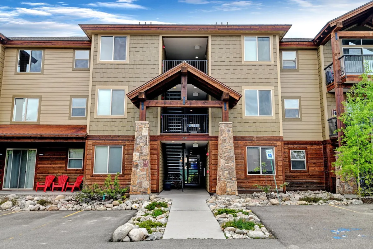 This exterior shot showcases the welcoming entrance to Trailhead Lodge Resort, featuring stone pillars, wooden accents, red chairs, and a landscaped pathway. People are gathering and relaxing outdoors, suggesting an inviting, community-oriented atmosphere.