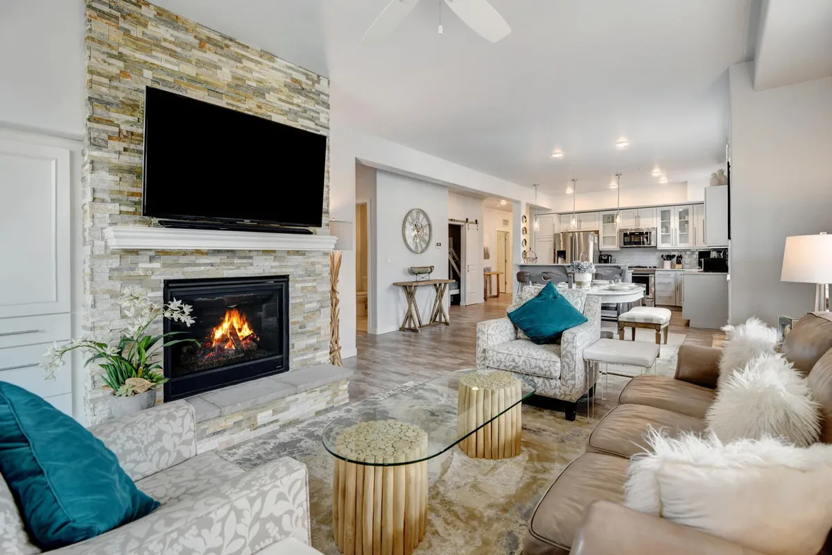A spacious, inviting living room featuring plush sofas gathered around a glowing stone fireplace, a large TV mounted above the mantle, and a sleek coffee table—perfect for gathering with family and friends. Floor-to-ceiling windows offer breathtaking sunset views, while warm lighting and mountain-inspired décor create a cozy, welcoming retreat.