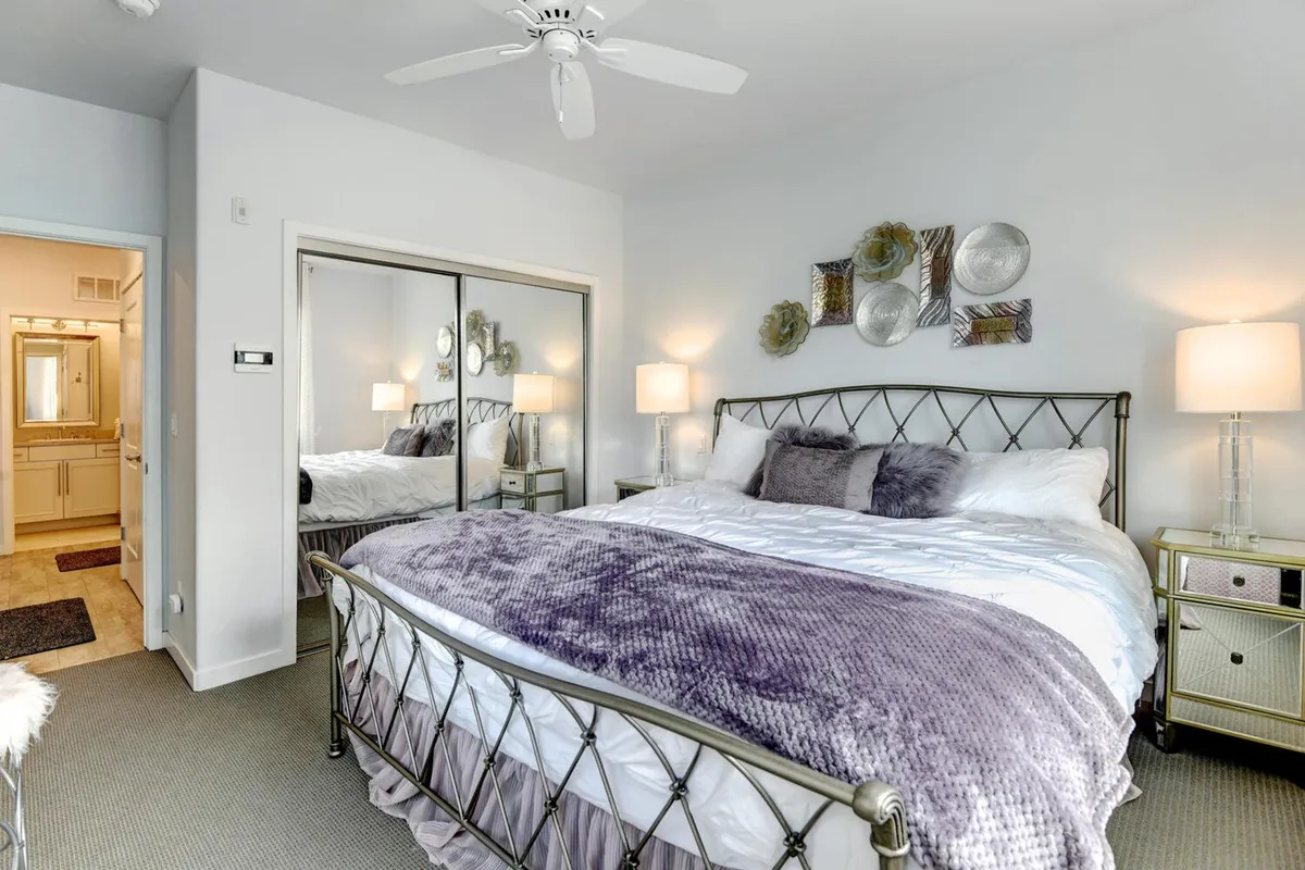 A queen bed topped with a purple throw blanket stands before a mirrored wardrobe and is flanked by nightstands with lamps. Decorative wall plates add character to this calm, cozy bedroom.