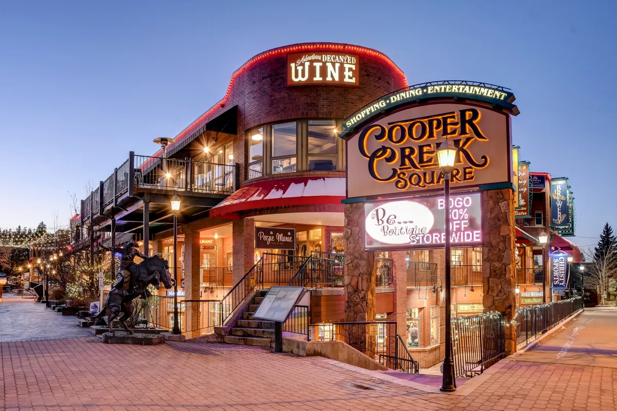 The image features the iconic Cooper Creek Square sign, a wine shop, and inviting outdoor seating areas, all illuminated by decorative lights. The scene is lively and welcoming, capturing the energetic spirit of downtown Winter Park.