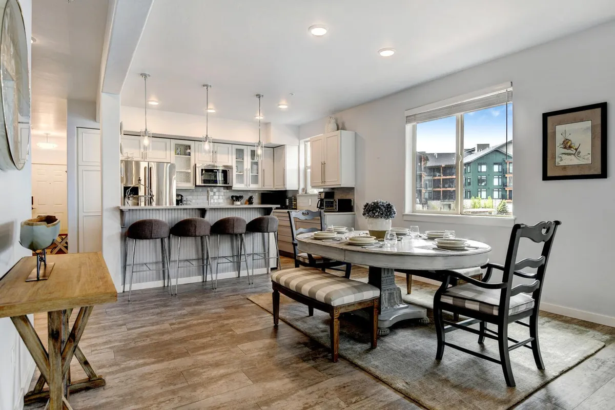 This image features a spacious and inviting dining area set with plates and cutlery, bar stools at the kitchen counter, premium kitchen appliances, and a window that lets in natural mountain light. The scene evokes a sense of togetherness, with ample space for group meals, cooking, and social activities.