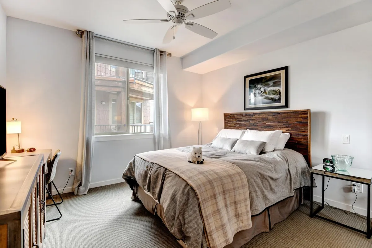 A king-sized bed with a wooden headboard sits beside a nightstand and lamp. Soft, sheer curtains filter natural light into the serene bedroom, creating an inviting space for rest and relaxation.
