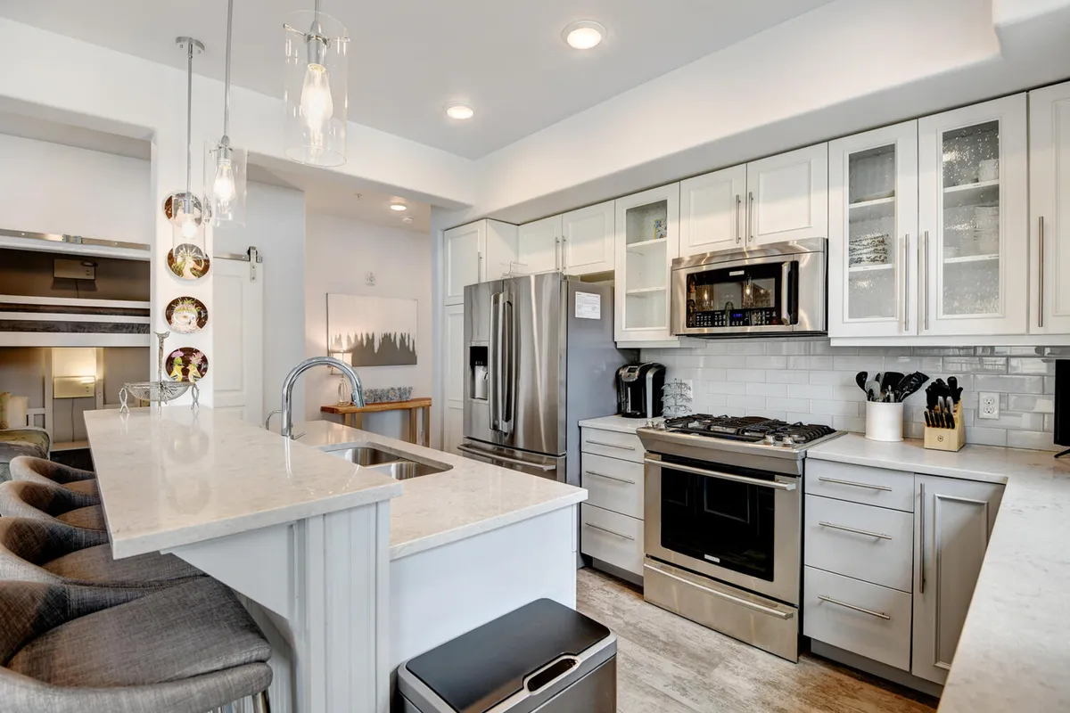 This perspective centers on the kitchen island with bar stools, stainless steel appliances, and decorative touches like hanging light fixtures and plates. The welcoming mood suggests both culinary creativity and connection, ideal for cooking together, snacks, or casual conversation.