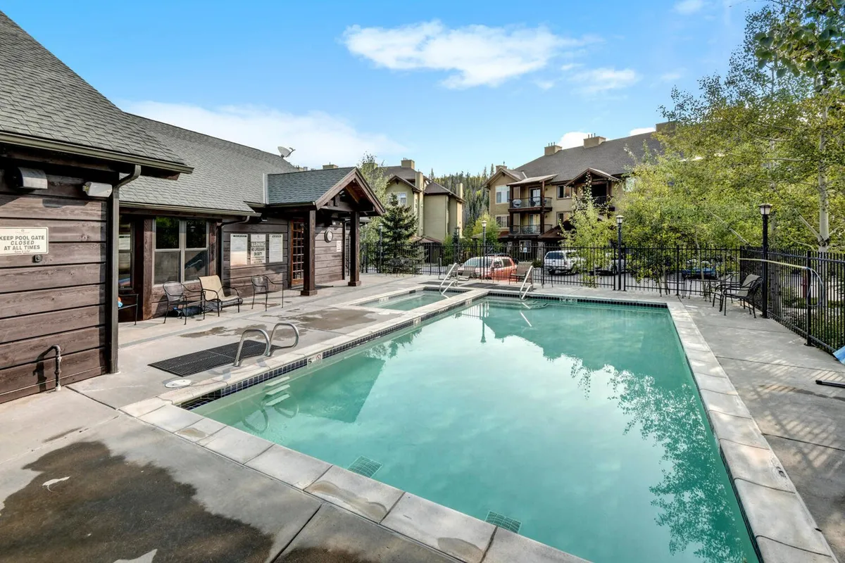 This image features a heated outdoor swimming pool surrounded by lounge chairs and a wooden clubhouse building, with lush trees in the background. The setting is inviting for swimming, sunbathing, and socializing, offering a relaxed, resort-style atmosphere.